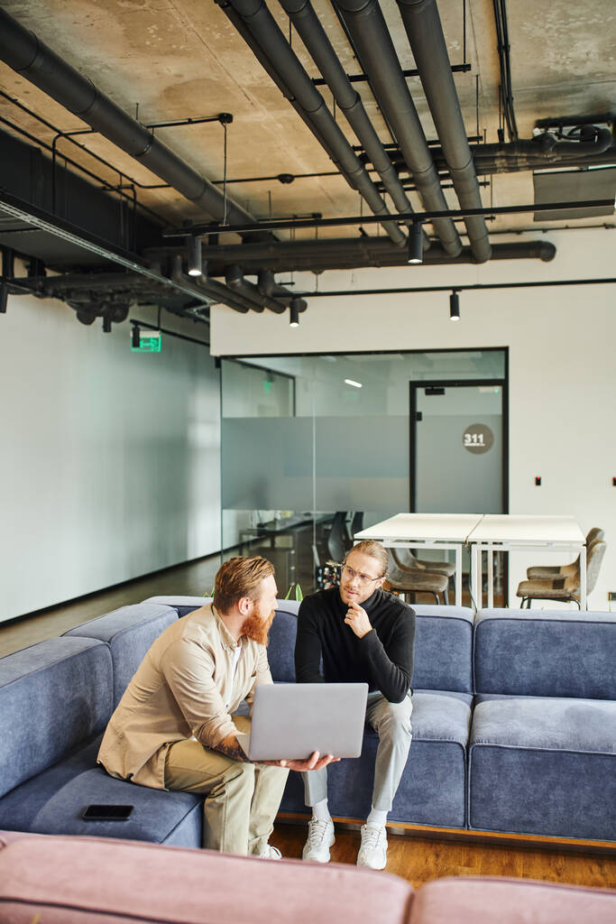 stylish business partners discussing startup project while sitting on comfortable couch with laptop in lounge of coworking office with high tech interior, business partnership concept - Photo, Image