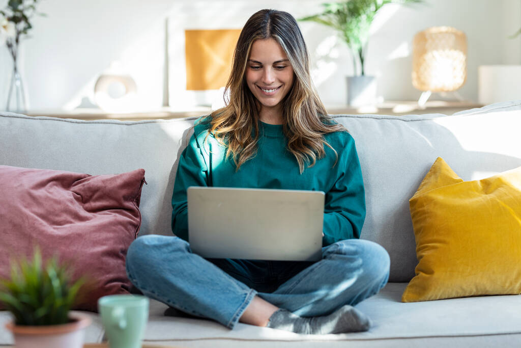 Aufnahme einer schönen freundlichen Frau, die mit Laptop arbeitet, während sie zu Hause auf der Couch im Wohnzimmer sitzt - Foto, Bild