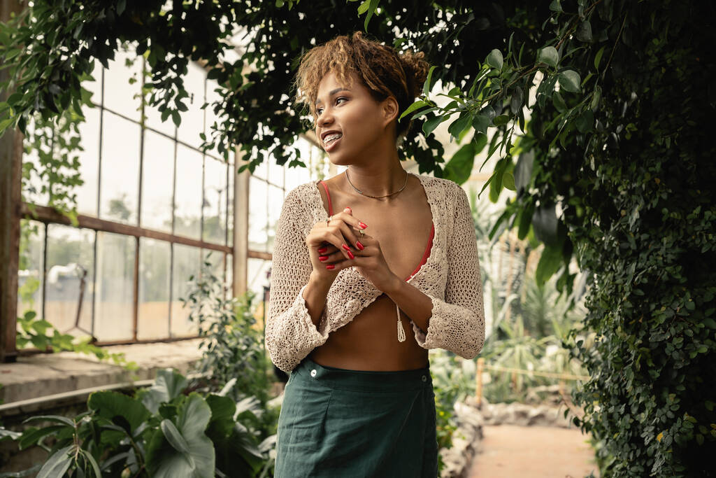 Pozitivní mladá africká Američanka v módní letní pletený top a sukně při pohledu pryč, zatímco stojí v blízkosti zelených rostlin v zahradním centru, stylová žena těší svěží tropické prostředí - Fotografie, Obrázek