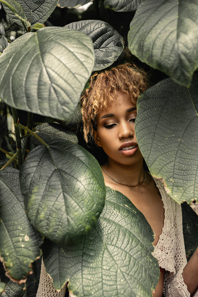 Trendy giovane donna afro-americana con il trucco indossando maglia superiore mentre in piedi vicino a foglie verdi sfocate in serra, donna elegante godendo di un ambiente tropicale lussureggiante - Foto, immagini