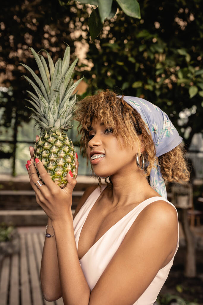 Portrait of smiling and stylish african american woman with braces in headscarf holding fresh pineapple and looking away while standing in blurred greenhouse, fashion-forward lady in tropical oasis - Photo, Image