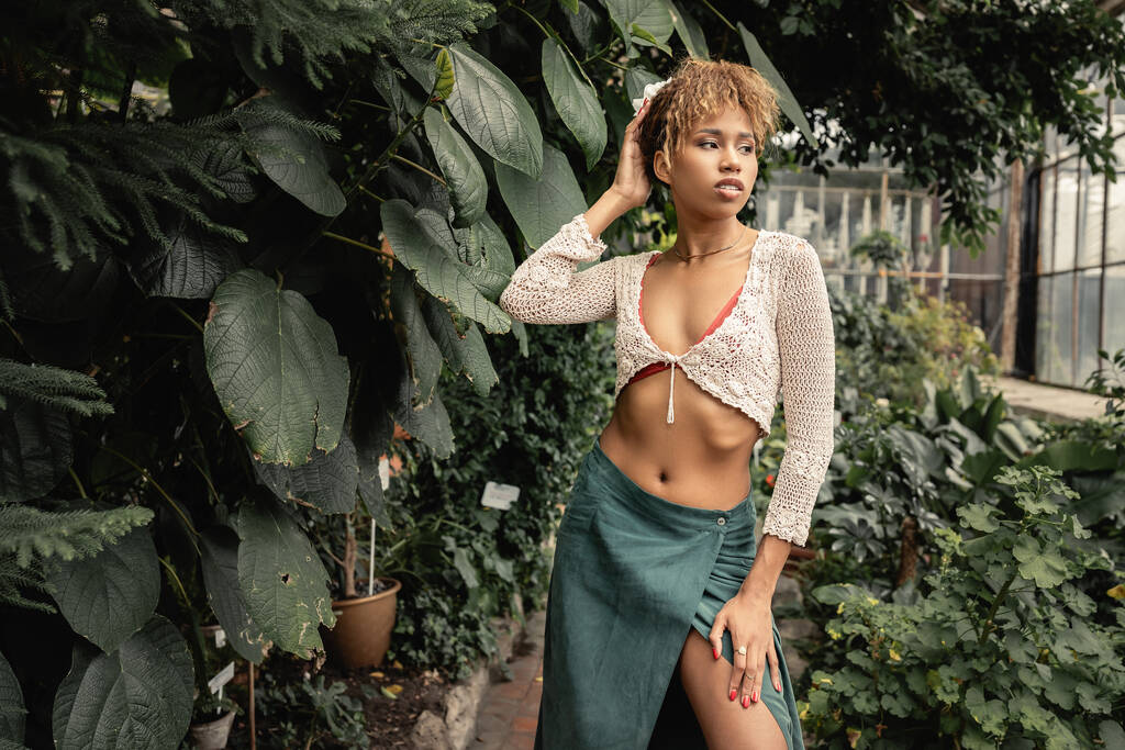 Μοντέρνα νεαρή Αφροαμερικανή με καλοκαιρινό ντύσιμο και πλεκτή μπλούζα που ποζάρει κοντά σε πράσινα φυτά και στέκεται σε θολό εσωτερικό κήπο, κομψή κυρία που περιβάλλεται από πλούσιο πράσινο, καλοκαίρι - Φωτογραφία, εικόνα