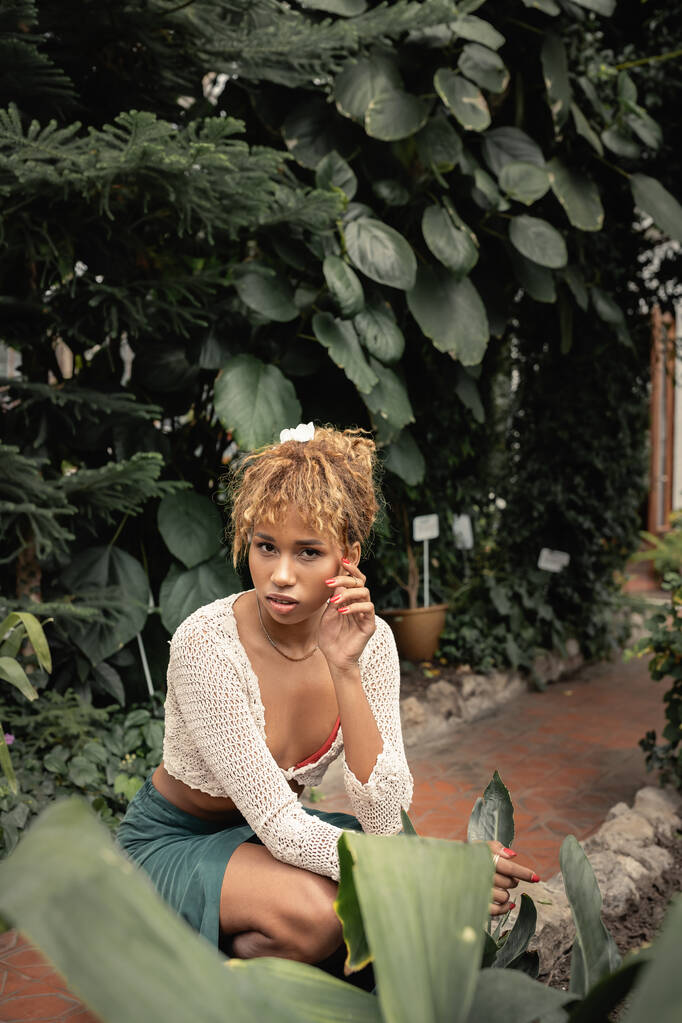 Divatos fiatal afro-amerikai nő nyári ruhában nézi a kamerát, miközben pózol közelében trópusi növények a háttérben narancs, elegáns hölgy körül buja növényzet, nyár - Fotó, kép