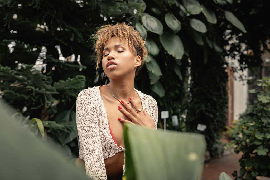 Genç ve şık Afro-Amerikan kadın yazın örgü örüyor göğsüne dokunuyor ve gözlerini kapatıyor arka planda kapalı bir bahçede dikiliyor, moda tutkunu tropikal flora ile kaynaşıyor. - Fotoğraf, Görsel