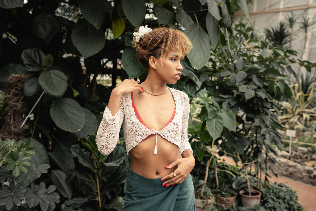 Elegante jovem mulher afro-americana em roupa de verão olhando para longe enquanto estava perto de plantas verdes no fundo no centro do jardim, fashionista misturando-se com flora tropical - Foto, Imagem
