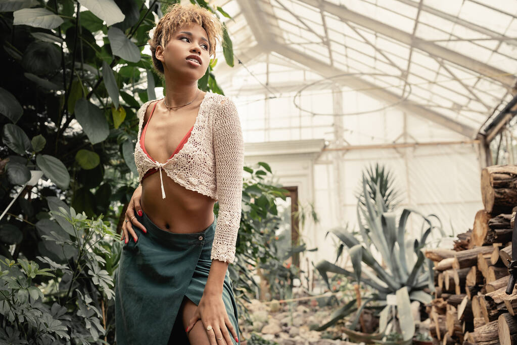 Модна молода афроамериканка в літньому вбранні, торкаючись стегна і дивлячись геть, стоячи в розмитому садовому центрі, стильна жінка з тропічним тлом - Фото, зображення