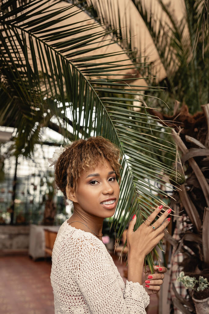 Usmívající se mladá africká Američanka s rovnátky v pleteném horním dotykovém brunch palmy a při pohledu na kameru v zahradním centru, módní pózování uprostřed tropické flóry, letní koncept - Fotografie, Obrázek