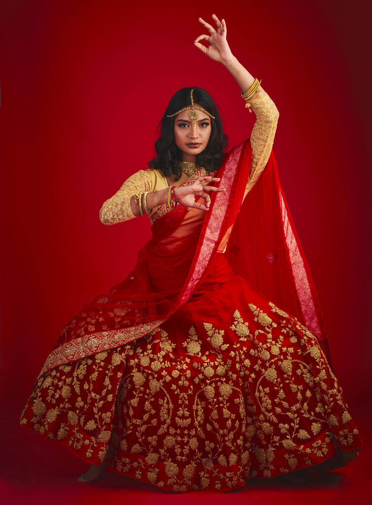 Πορτρέτο, χορός και Ινδή γυναίκα με μόδα, παραδοσιακό φόρεμα και γιορτή σε ένα κόκκινο φόντο στούντιο. Πρόσωπο, θηλυκό πρόσωπο ή μοντέλο με πολιτιστικά ρούχα, κοσμήματα και χορό με ομορφιά. - Φωτογραφία, εικόνα