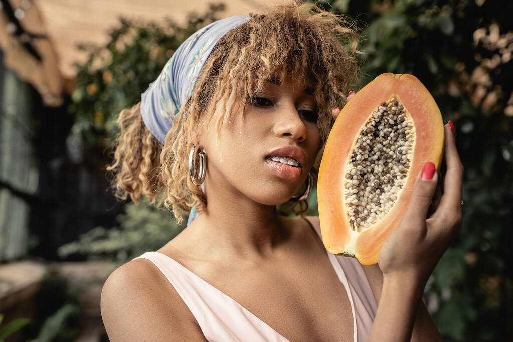 Portrait of trendy young african american woman in headscarf and summer outfit holding cut and ripe papaya while standing in blurred garden center, stylish lady blending fashion and nature - Photo, Image