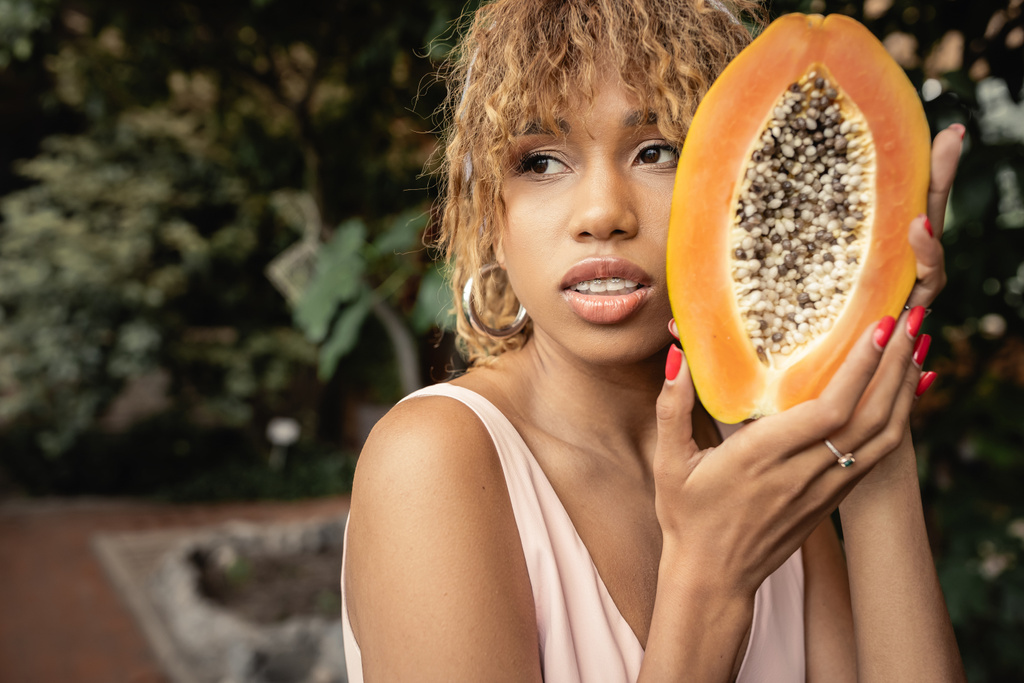 Confident young african american woman with braces and summer outfit holding fresh and ripe papaya and looking away in blurred greenhouse, stylish lady blending fashion and nature, summer concept - Photo, Image