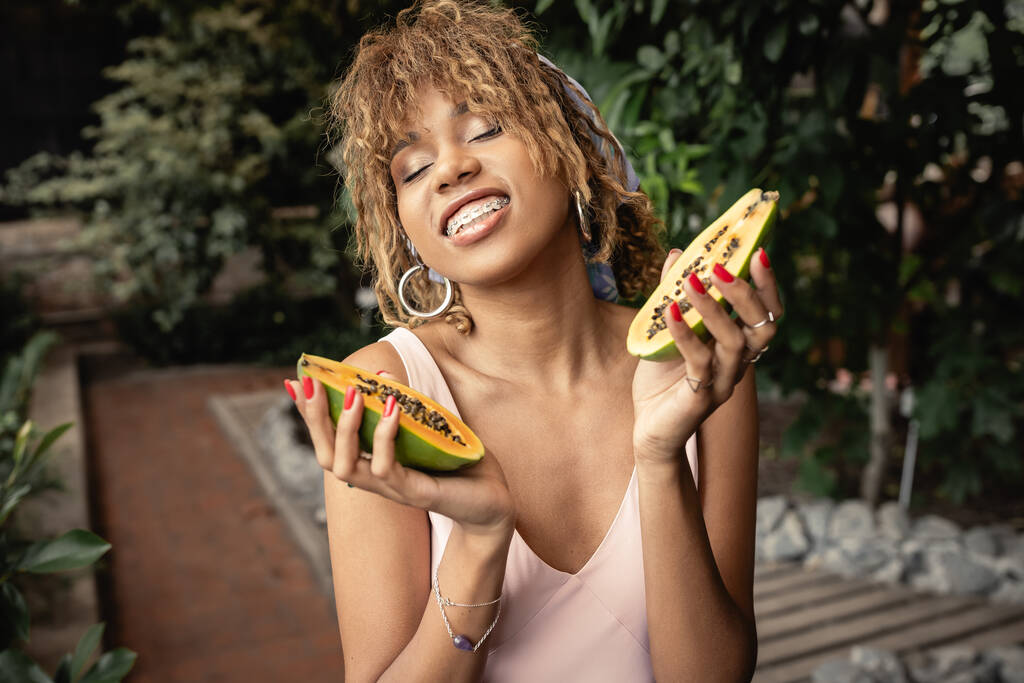 Mujer joven afroamericana sonriente con frenos que combaten el traje de verano y cierran los ojos mientras sostiene la papaya fresca en el jardín interior borroso, dama de moda inspirada en las plantas tropicales - Foto, imagen