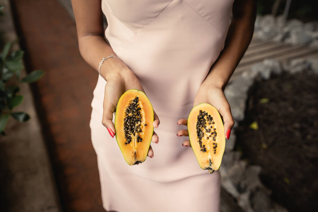 Vista recortada de la joven afroamericana de moda en vestido de verano sosteniendo papaya recién cortada mientras está de pie en el centro del jardín, señora de la moda hacia adelante inspirada en las plantas tropicales, concepto de verano - Foto, imagen