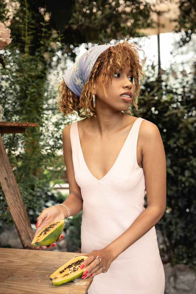 Mladá africká Američanka ve stylovém šátku a letních šatech držící čerstvou papáju a odvracející se v rozmazaném skleníku, módní dáma inspirovaná tropickými rostlinami, letní koncept - Fotografie, Obrázek