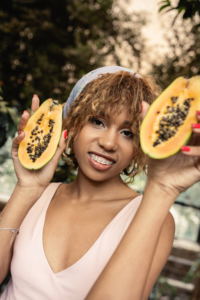 Cheerful young african american woman with braces wearing summer dress and headscarf while holding cut papaya and looking at camera in orangery, fashion-forward lady inspired by tropical plants - Photo, Image
