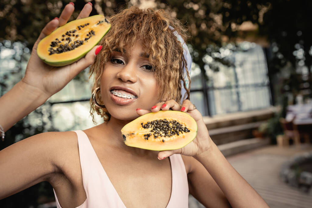 Portré pozitív fiatal afro-amerikai nő fogszabályzó visel nyári ruhát, és gazdaság friss papaya miközben áll a narancs, trendi nő körül trópusi buja - Fotó, kép