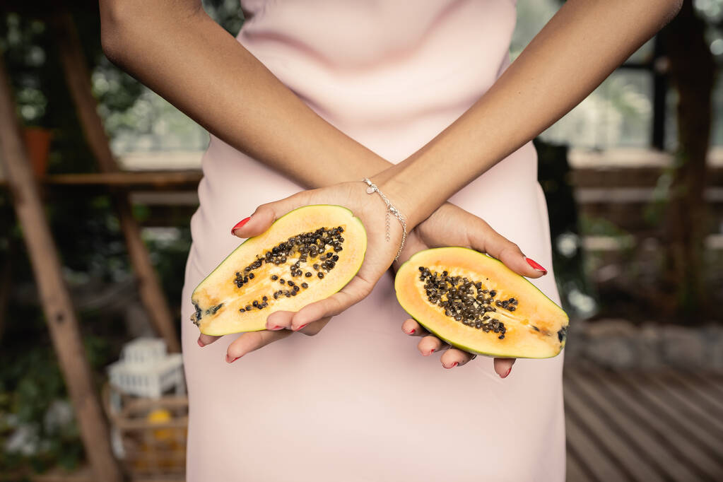 Vue recadrée de la jeune femme afro-américaine en robe d'été tenant la papaye fraîche et debout dans un jardin intérieur flou, femme à la mode entourée de luxuriance tropicale - Photo, image