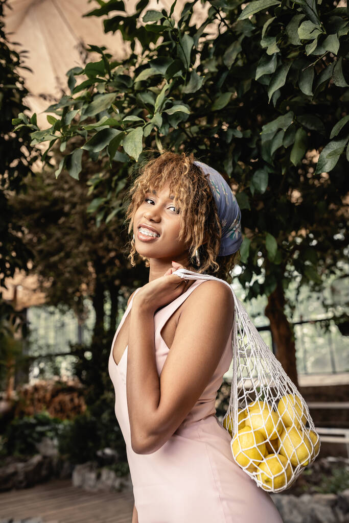 Portret van een lachende Afro-Amerikaanse vrouw met beugels in zomerjurk met mesh bag met verse citroenen en kijkend naar de camera in de binnentuin, stijlvolle dame die geniet van tropische sfeer - Foto, afbeelding
