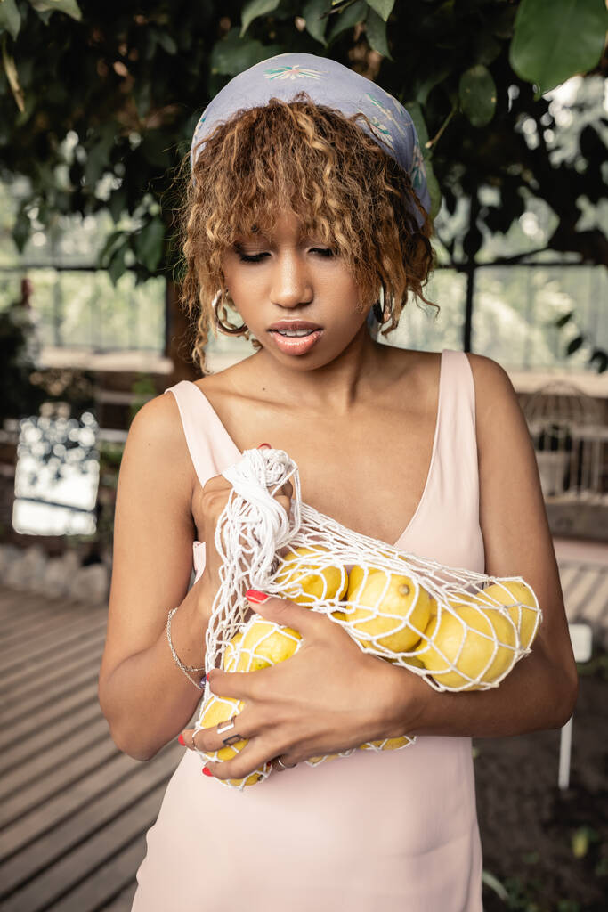 Retrato de una joven afroamericana con pañuelo en la cabeza y vestido de verano sosteniendo bolsa de malla con jugosos limones y de pie en un borroso naranjero de fondo, elegante dama disfrutando de la atmósfera tropical - Foto, Imagen