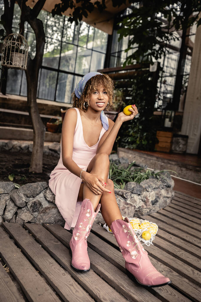 Full length of smiling young african american woman in boots and summer outfit holding fresh lemon while sitting near mesh bag in blurred orangery, chic woman in tropical garden, summer concept - Photo, Image