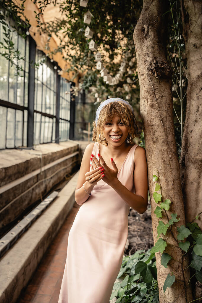 Joyeux et élégant jeune femme afro-américaine avec bretelles en foulard et robe d'été en regardant la caméra tout en se tenant près des arbres en orangerie, femme élégante avec des plantes tropicales en toile de fond - Photo, image
