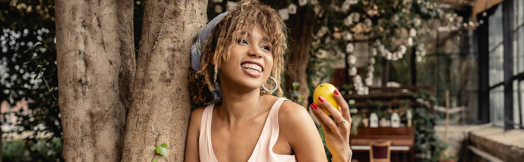 Mujer afroamericana joven y de moda con tirantes y ropa de verano sosteniendo limón maduro y de pie cerca de los árboles en el centro del jardín, señora de la moda hacia adelante en armonía con la flora tropical, pancarta  - Foto, Imagen