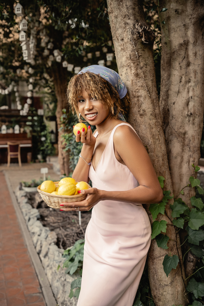 Moderna mujer afroamericana joven en traje de verano sonriendo y sosteniendo cesta con limones cerca de los árboles en el jardín interior borroso, señora de la moda hacia adelante en armonía con la flora tropical, concepto de verano - Foto, imagen