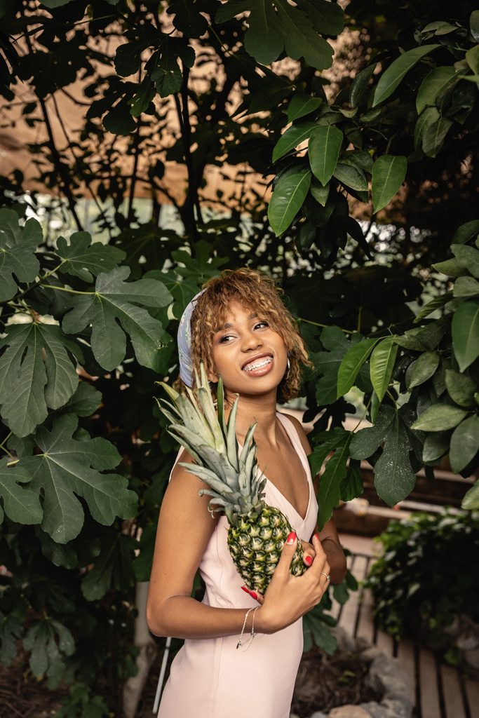 Smiling young african american woman with braces wearing summer outfit and holding pineapple and standing near plants in orangery, woman in summer outfit posing near lush tropical plants - Photo, Image