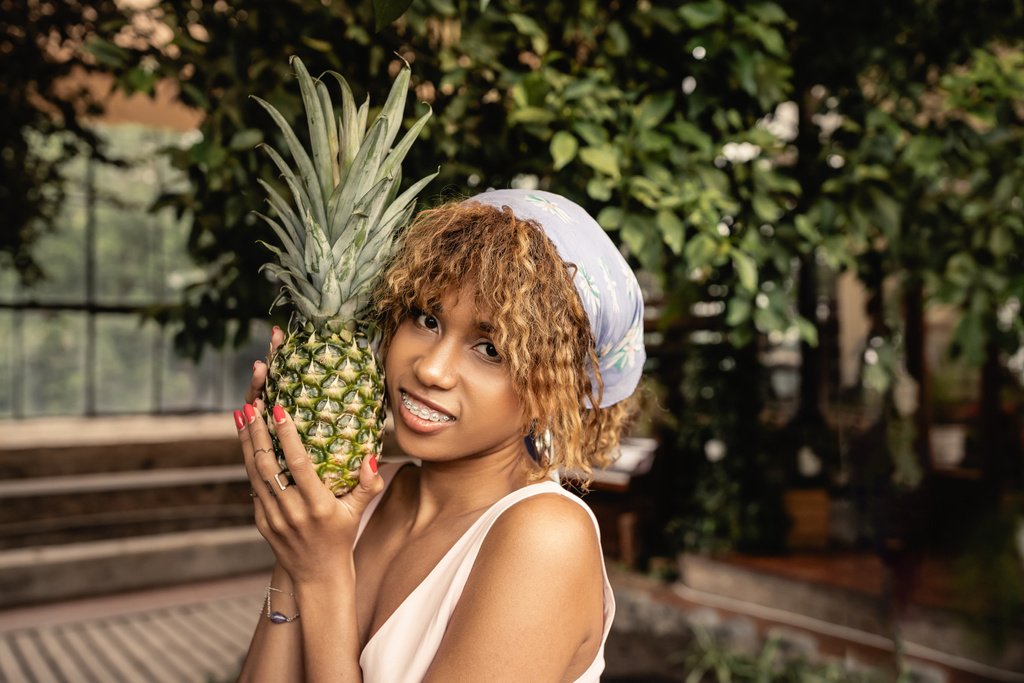 Glimlachende jonge Afro-Amerikaanse vrouw met beugel en hoofddoek met verse ananas en kijkend naar de camera in wazige oranjerie, stijlvolle vrouw in zomerkleding omgeven door tropisch gebladerte - Foto, afbeelding