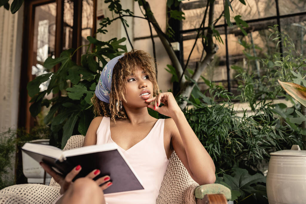 Jonge en zelfverzekerde Afrikaans-Amerikaanse vrouw in zomerse outfit met boek en zittend op een fauteuil in wazige binnentuin, stijlvolle vrouw in zomerse outfit omringd door tropisch gebladerte - Foto, afbeelding