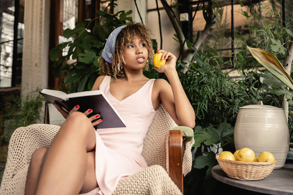 Mujer afroamericana joven de moda en traje de verano sosteniendo limón fresco y sosteniendo libro en sillón en el centro del jardín, mujer elegante usando traje de verano rodeado de follaje tropical - Foto, Imagen