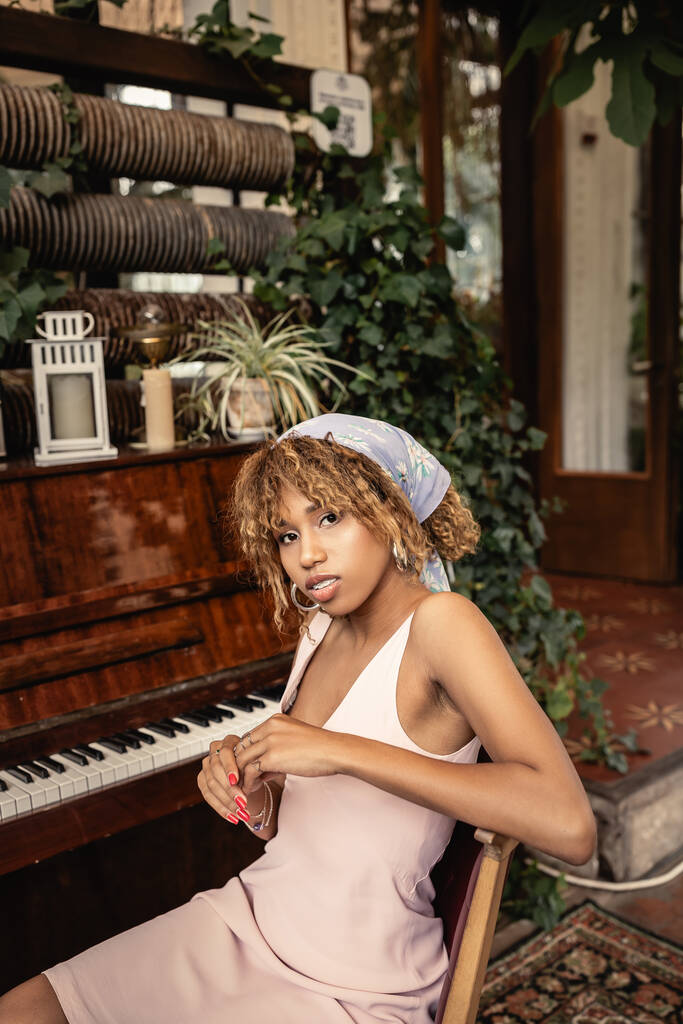 Modern young african american woman in summer dress looking at camera while sitting near piano and blurred plants in orangery, woman in comfortable and trendy summer outfit - Photo, Image
