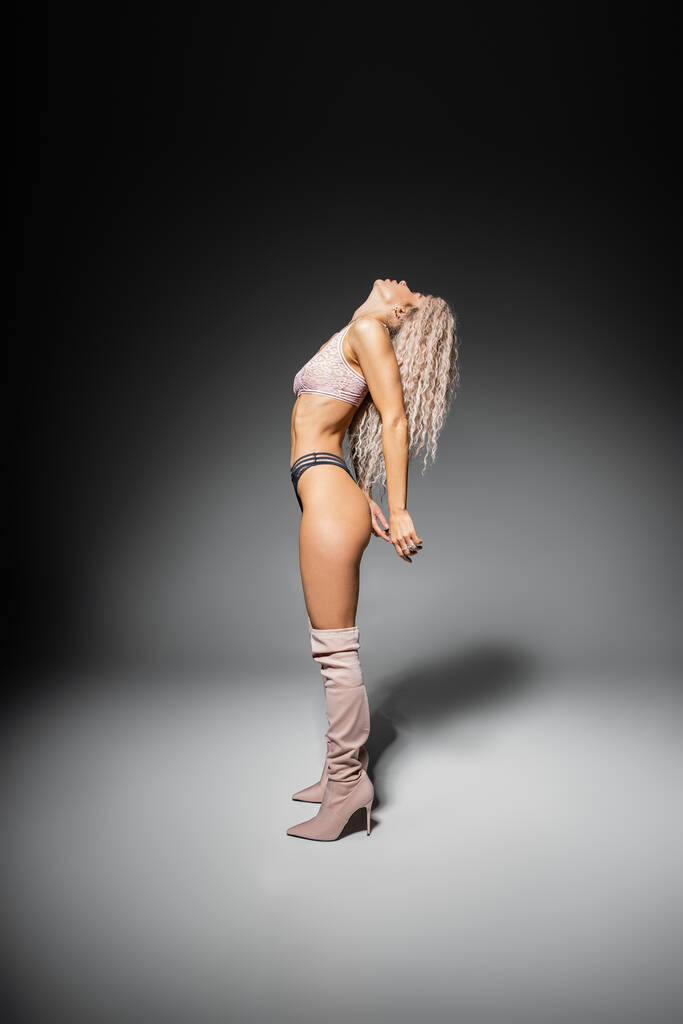 sexualité et de la mode, l'expression de soi moderne, toute la longueur du modèle féminin avec le corps tonique et ondulé frêne cheveux blonds posant en dentelle sous-vêtements et bottes élégantes hautes sur fond gris, vue de côté - Photo, image