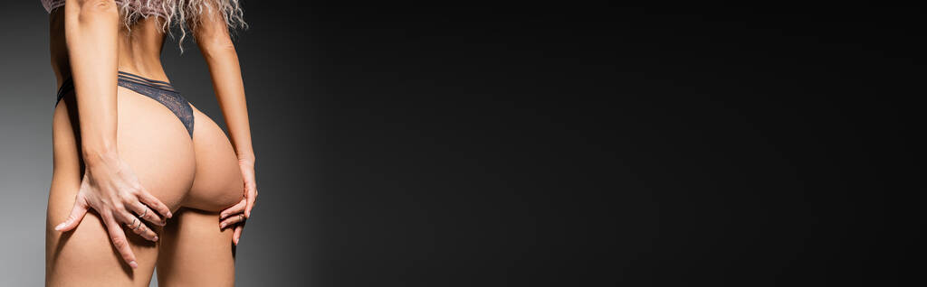 sexualidade e moda, vista cortada de mulher provocante com corpo tonificado tocando sexy nádegas enquanto posando em calcinhas de renda em fundo preto e cinza, banner com espaço de cópia - Foto, Imagem