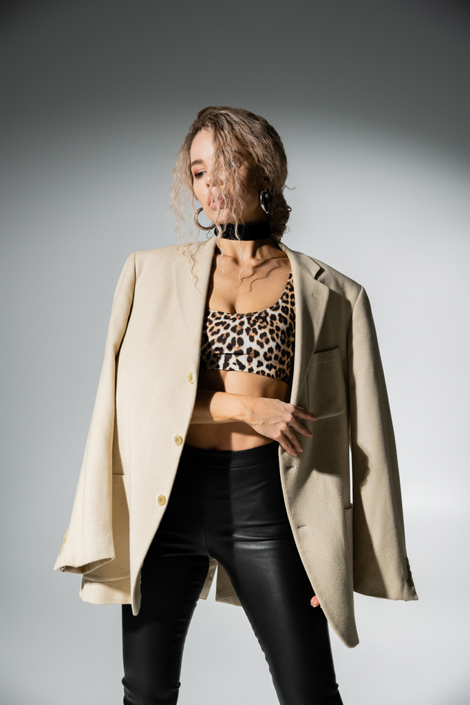 sensuality and fashion, charming woman with dyed ash blonde hair and slender body posing in animal print crop top and black latex pants, with beige blazer on shoulders on grey background - Photo, Image