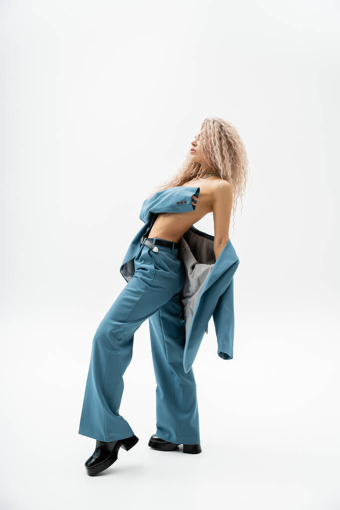 longitud completa de la mujer expresiva y de moda que cubre el pecho desnudo con el brazo mientras posa en traje azul de gran tamaño sobre el cuerpo sin camisa sobre fondo gris, estilo glamour, vista lateral - Foto, imagen