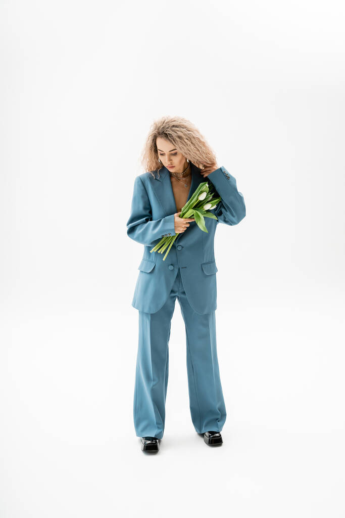 vrouwelijkheid en stijl, volledige lengte van stijlvolle en mooie vrouw met geverfd as blond haar met boeket van witte tulpen terwijl staan in blauw oversize pak op grijze achtergrond - Foto, afbeelding