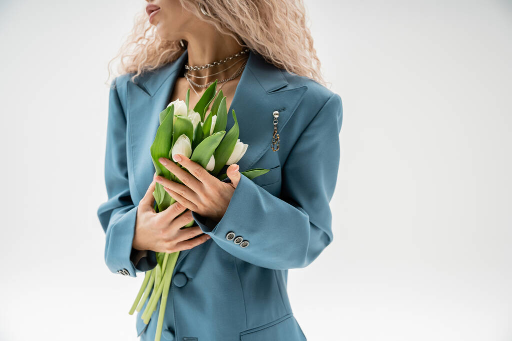 partial view of fashionable woman in blue oversize blazer holding bouquet of white tulips while standing on grey background, wavy ash blonde hair, silver accessories, modern style - Photo, Image