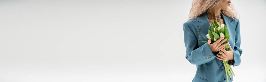 vista ritagliata di donna alla moda con capelli biondi cenere ondulati con bouquet di tulipani bianchi mentre in piedi in tuta oversize blu su sfondo grigio, banner con spazio copia, sensualità e moda - Foto, immagini