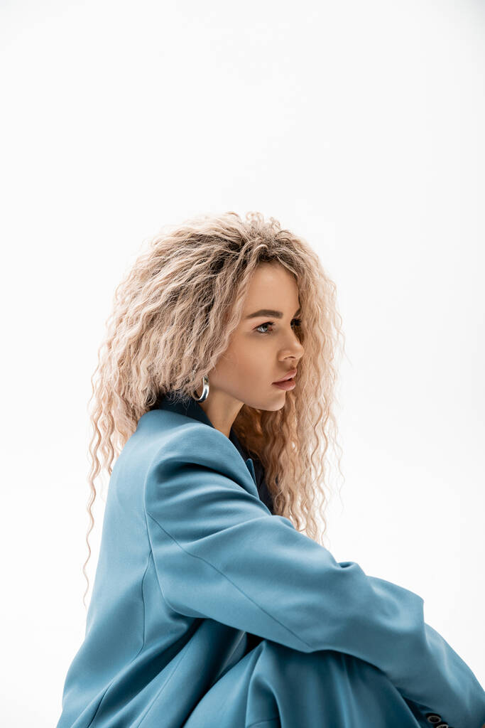 portrait de profil de femme envoûtante avec des cheveux blonds cendrés ondulés portant blazer élégant bleu et regardant loin sur fond gris, charmant et sensuel modèle féminin, la mode moderne - Photo, image
