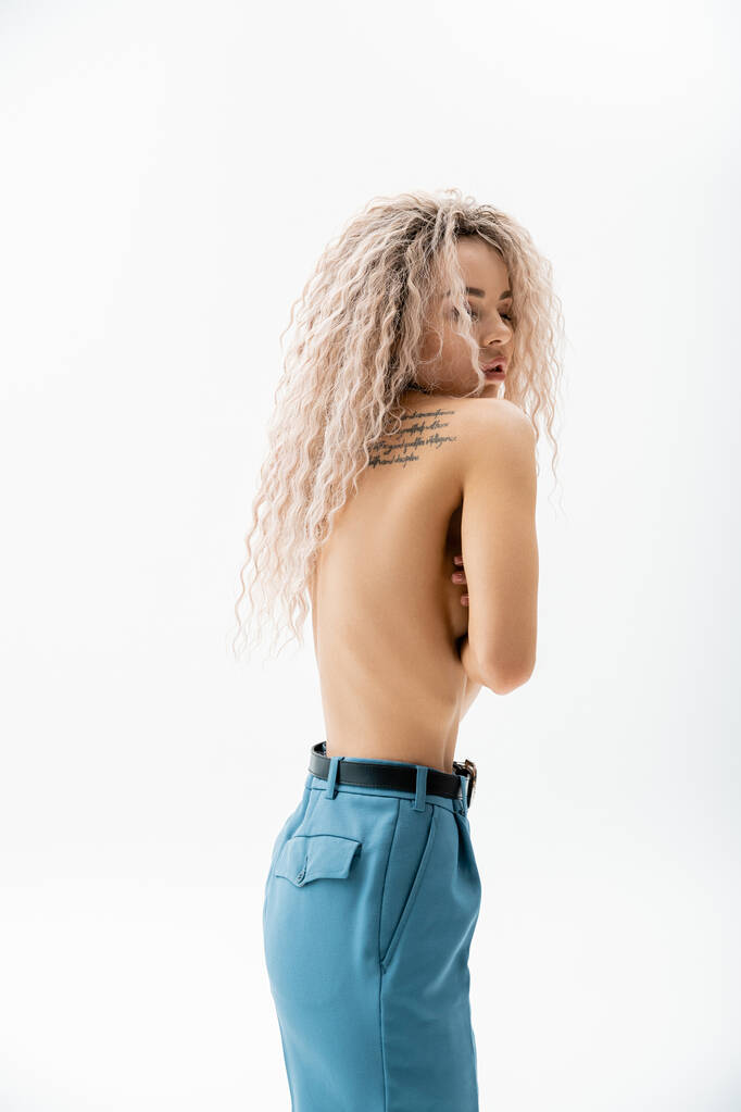 seductive and expressive woman with shirtless tattooed body obscuring naked breast with hands while standing in blue oversize pants on grey background, fashion and sexuality, modern individuality - Photo, Image