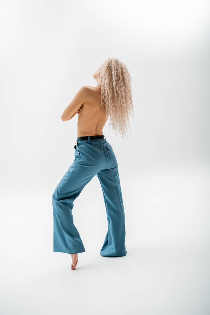 plná délka sexy, bez trička a bosá žena s barveným popelem blond vlasy pózující v modré oversize kalhoty na šedém pozadí, štíhlé tělo, individualita a self-výraz - Fotografie, Obrázek
