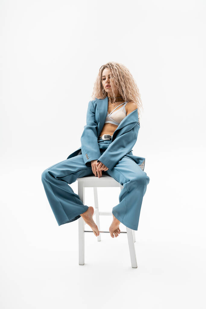 full length of sexy, appealing and trendy barefoot woman sitting on chair in bra and blue oversize suit on grey background, wavy ash blonde hair, sensual individuality, modern fashion - Photo, Image