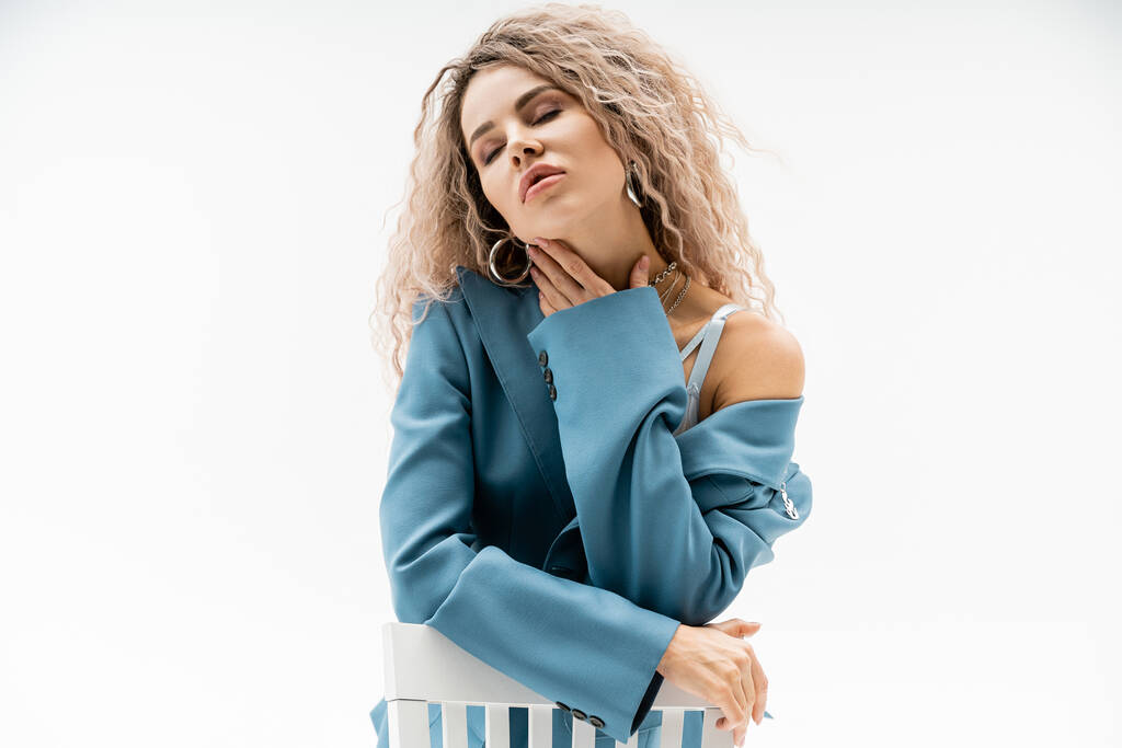 seductive and beautiful woman with wavy ash blonde hair and closed eyes touching neck and sitting on chair in blue oversize blazer and silver accessories on grey background - Photo, Image