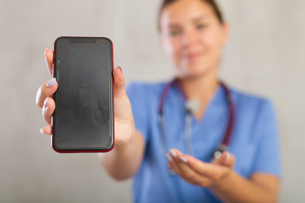 Mujer joven positiva en uniforme médico posando en estudio con teléfono móvil - Foto, Imagen