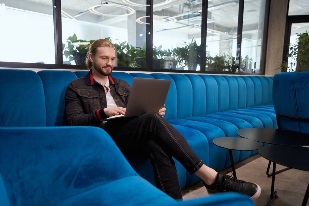 Красивый молодой человек сидит на синем диване в зоне отдыха, он в чате онлайн - Фото, изображение