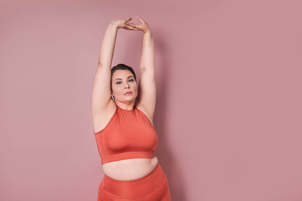 Mujer de talla grande satisfecha en ropa de fitness posando en el estudio con fondo rosa. Mujer seria en ropa deportiva sonriendo después de la sesión de entrenamiento - Foto, imagen