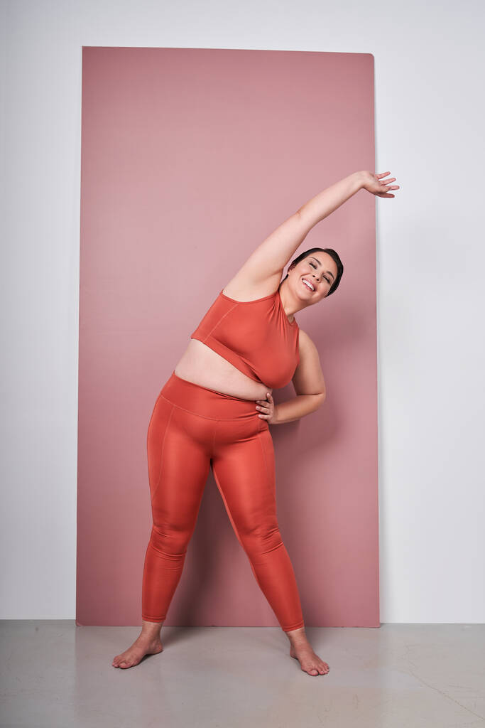スタジオピンクの背景に孤立したフィット感の運動を行うスポーツウェアを身に着けている笑顔脂肪太りすぎの女性のフル長さの写真。ワークアウトスポーツ、フィットネス、ボディポジティブなコンセプト - 写真・画像