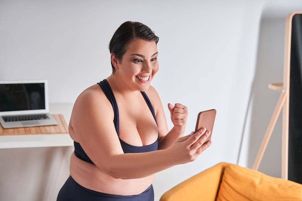 La bloguera deportiva positiva para el cuerpo se siente alegre mientras habla de abdominales con sus suscriptores. Mujer feliz usando smartphone - Foto, imagen
