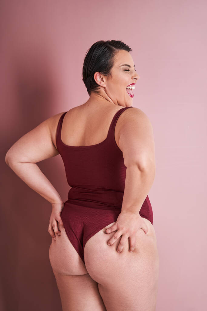 Rückenansicht der überglücklichen Emotionalkörper-positiven Frau, die ihren natürlichen Körper genießt, während sie vor rotem Studiohintergrund steht - Foto, Bild
