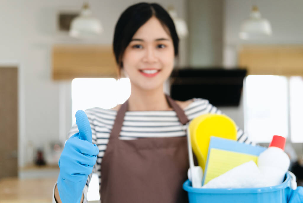 Uśmiechnięta młoda kobieta gospodyni domowa w brązowym fartuchu i niebieskich rękawiczkach trzymając butelki i pokazując kciuk w górę, Stojąc w nowoczesnej kuchni, profesjonalne sprzątanie. - Zdjęcie, obraz